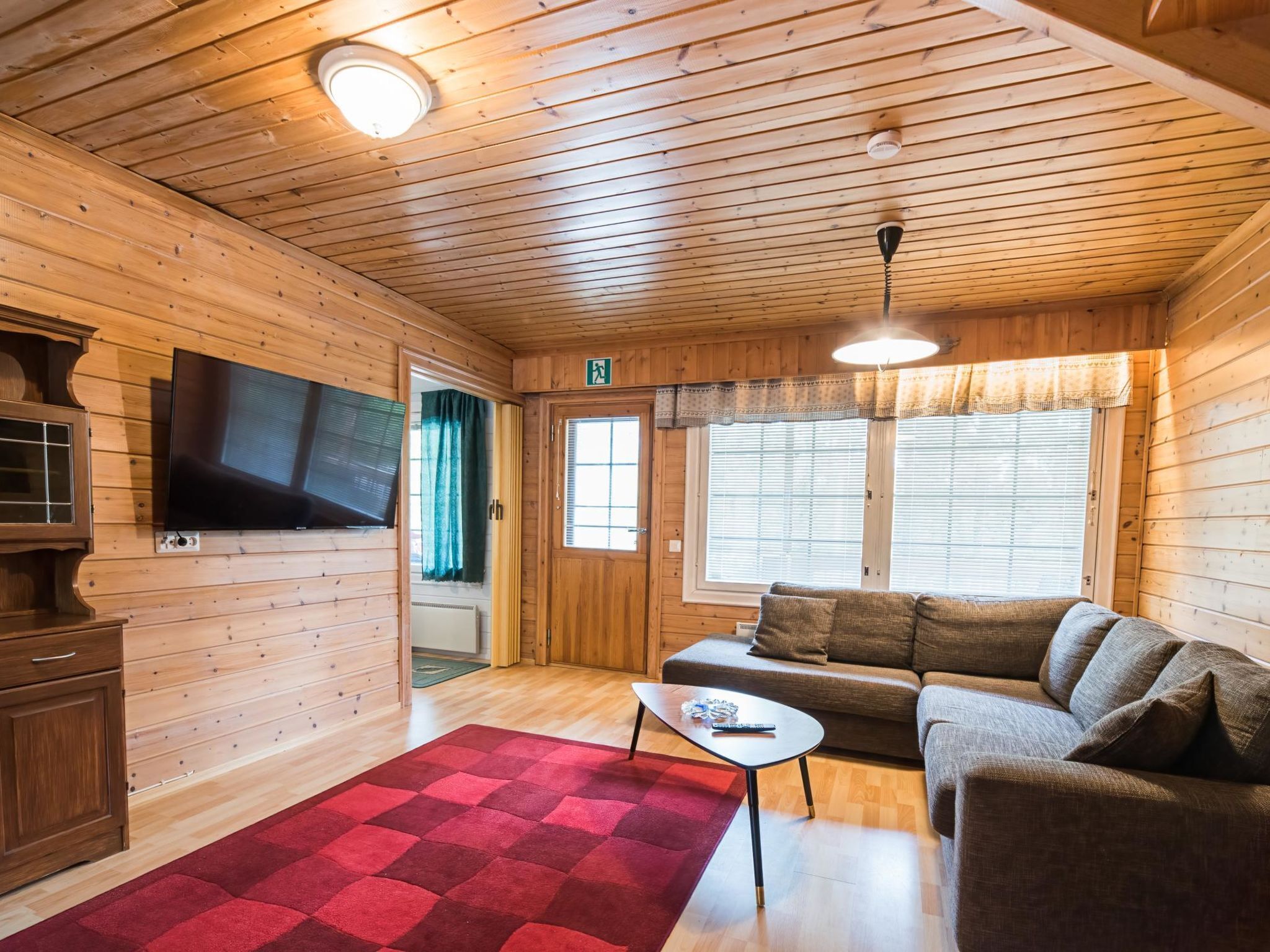 A cozy cabin interior with a wooden ceiling, a television mounted on the wall, a sofa, a small table, and a red rug.