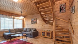 Un intérieur chaleureux avec des éléments en bois, comprenant un canapé, une table basse et un tapis rouge. Un escalier en bois mène à un niveau supérieur.
