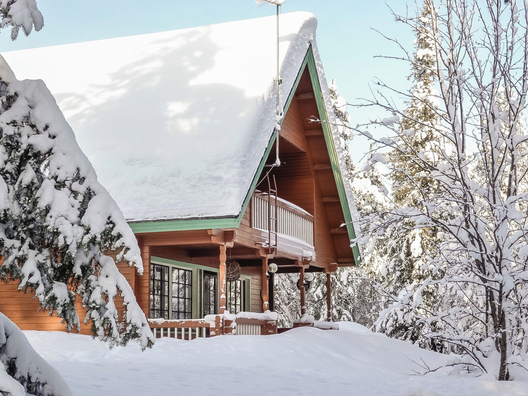 A wooden cabin with a snow-covered roof, a balcony, and a porch surrounded by snow-laden trees.