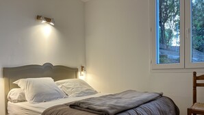 Een netjes opgemaakt bed met hoofdbord, een bedlampje en een raam met uitzicht op bomen.