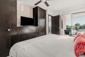 A bedroom with a bed, a TV on a wooden cabinet, and a balcony with a view of trees.