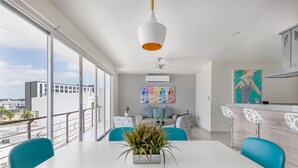 A modern living room with a white table, teal chairs, and a large window offering a city view. - Luxury 2 Bedroom Apartments (Cancun)