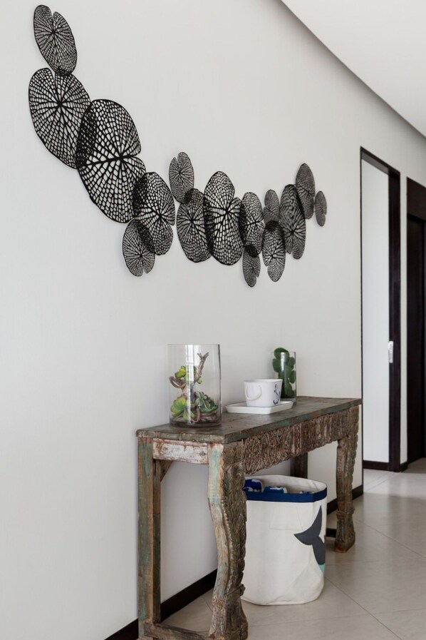 Un couloir orné d’une décoration murale représentant des motifs noirs en forme de feuilles, une table en bois rustique sur laquelle sont posés un vase en verre et une tasse, ainsi qu’une poubelle décorée d’un motif de baleine.