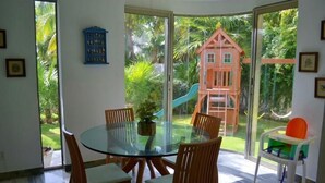 A dining area with a glass table and wooden chairs, a playhouse with a slide, and a view of a lush garden through large glass doors.
