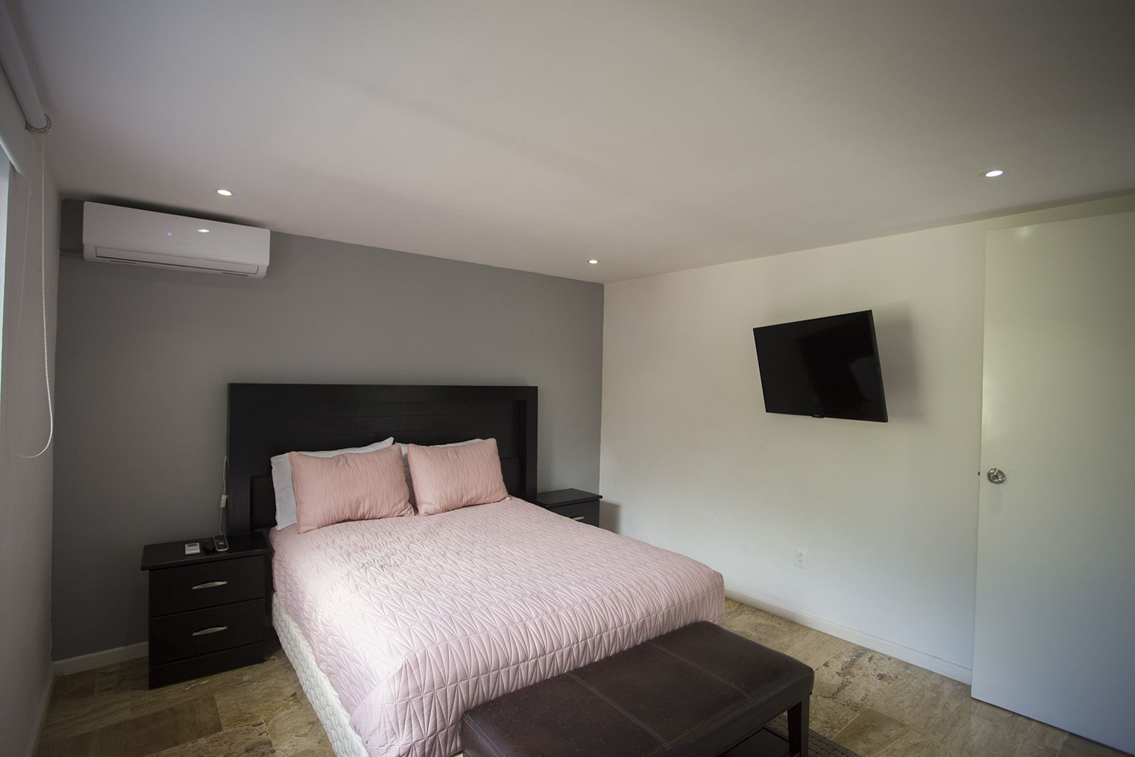 A bedroom with a bed, a nightstand, a bench, a TV, and an air conditioning unit.