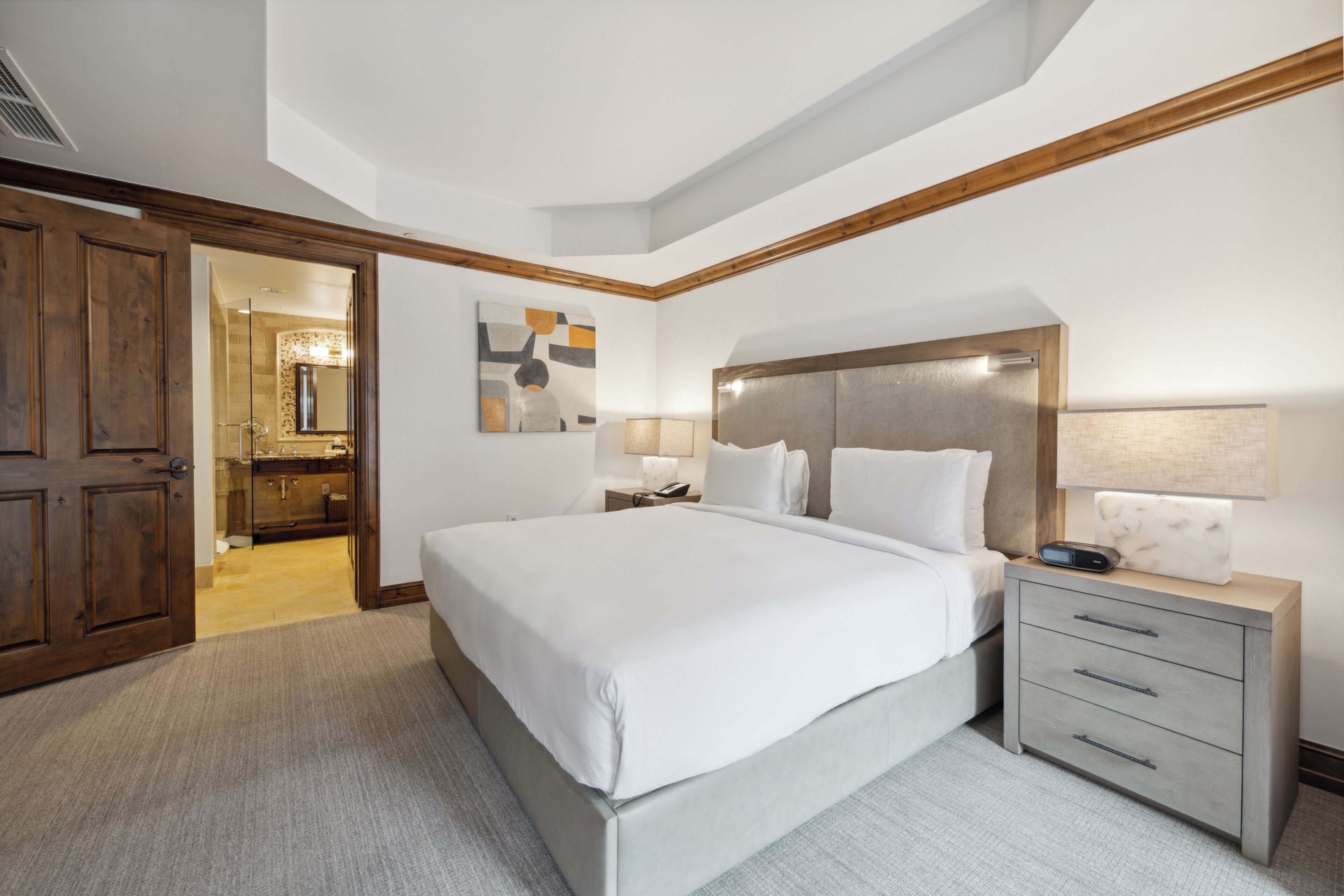 Una habitación de hotel moderna con una cama grande, una mesita de noche con lámpara y vistas al baño.