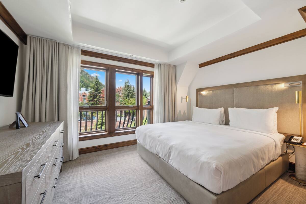 A hotel room with a large bed, a desk with drawers, a telephone, and a window with curtains.