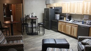 Une cuisine compacte avec des meubles en bois, un réfrigérateur, un four à micro-ondes, une cuisinière et une machine à café. On y trouve une table à manger avec des chaises et un pouf à motifs.