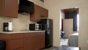Une cuisine compacte équipée d’un four à micro-ondes, d’un réfrigérateur et d’une machine à café. On y trouve une petite table avec une télécommande. Une porte donne accès à une chambre à coucher avec un lit et une chaise.