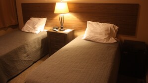 A hotel room with two single beds, a nightstand with a lamp, a remote control, and a small table.