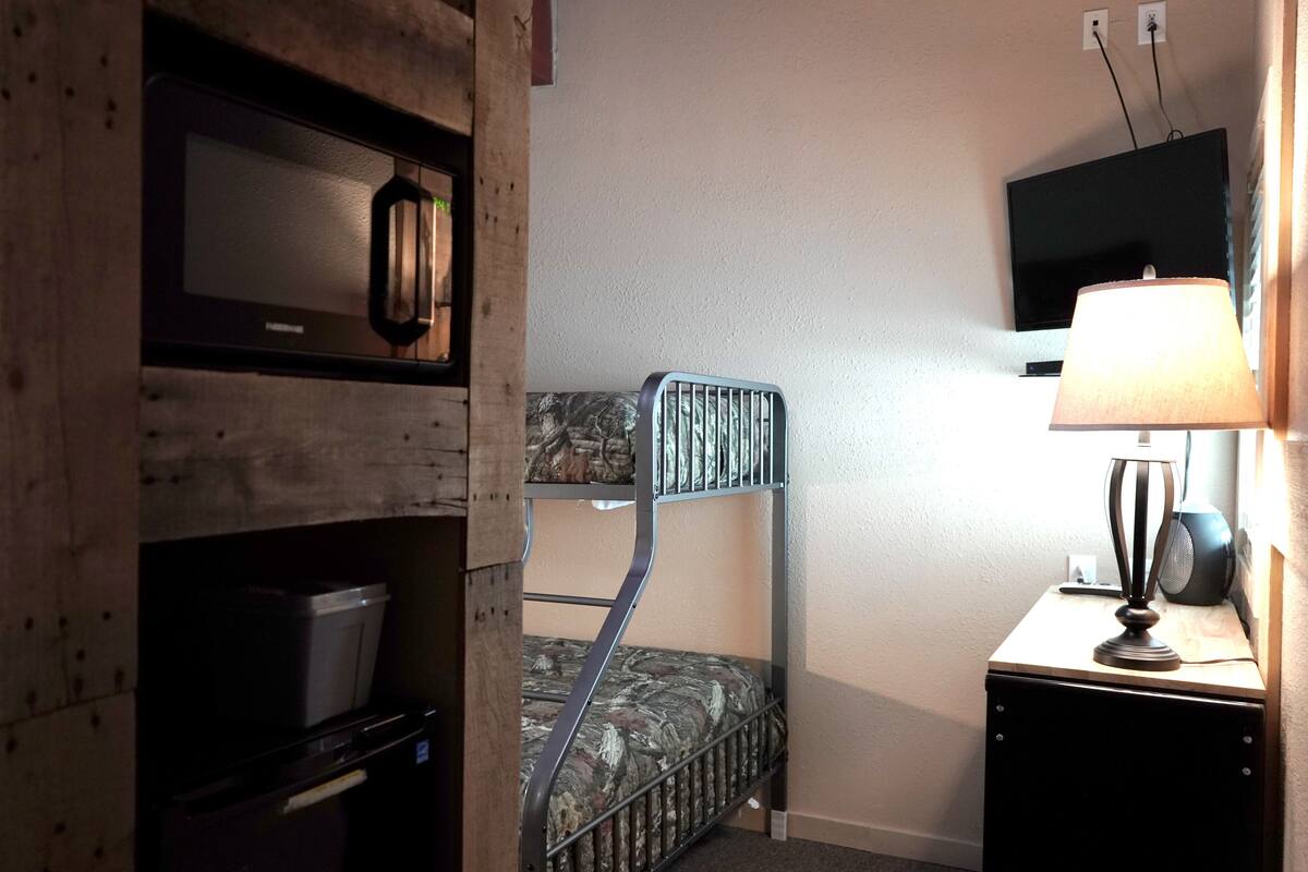 Un lit superposé avec une couette à motifs camouflage, un four à micro-ondes, une télévision et une lampe sur une table de chevet.