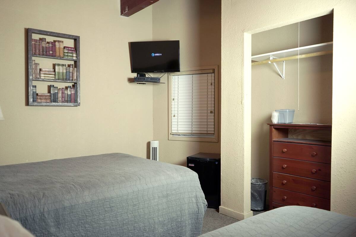 Une chambre d’hôtel avec deux lits, un téléviseur fixé au mur, une bibliothèque, un radiateur, une fenêtre avec des stores et une commode en bois.