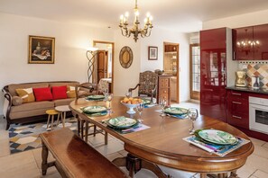 Un coin repas avec une table en bois dressée pour quatre personnes, un canapé avec des coussins rouges, un lustre et une cuisine avec des meubles rouges.