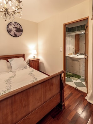 A bedroom with a wooden bed, a nightstand with a lamp, a chandelier, a framed picture on the wall, and a view into the bathroom through an open door.
