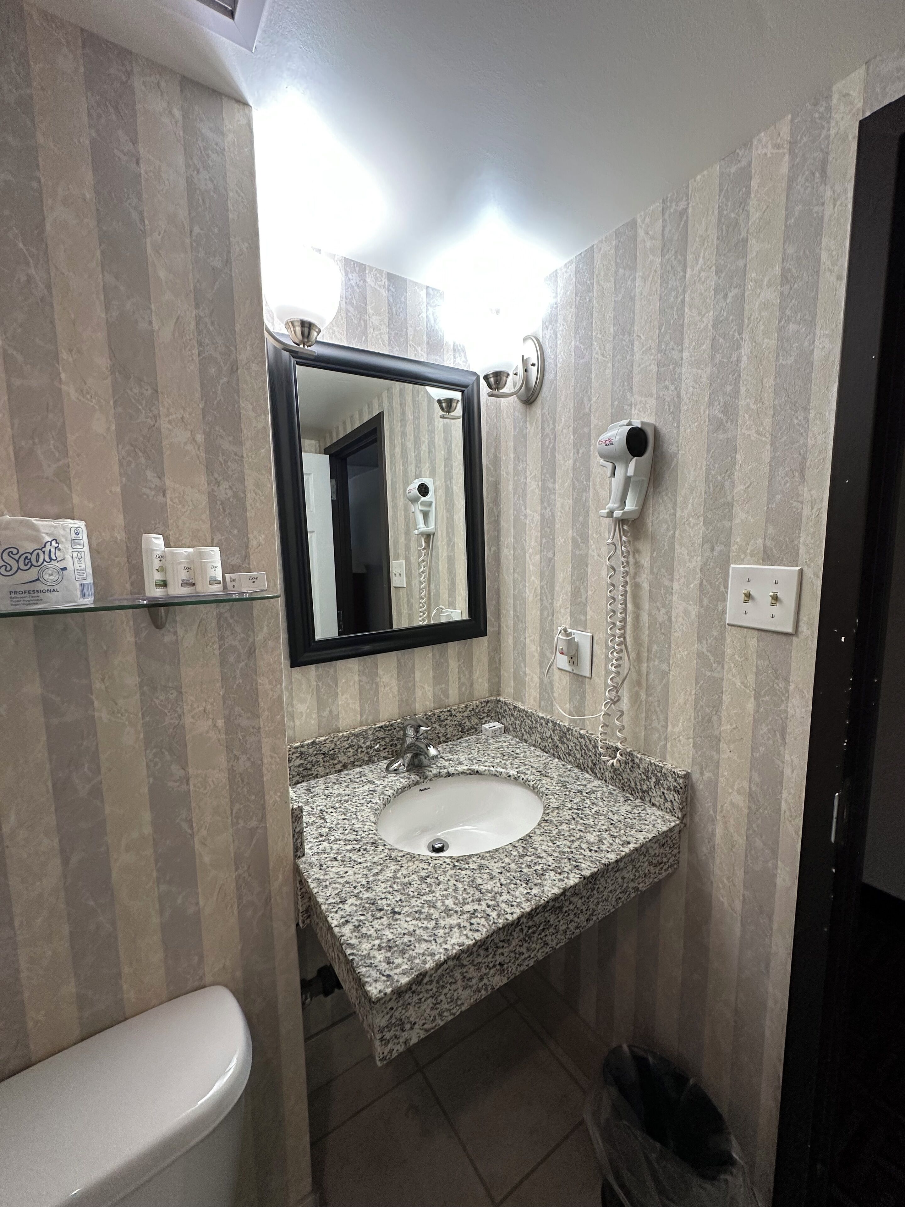 A bathroom with a granite sink, a toilet, a mirror, and a hairdryer mounted on the wall.