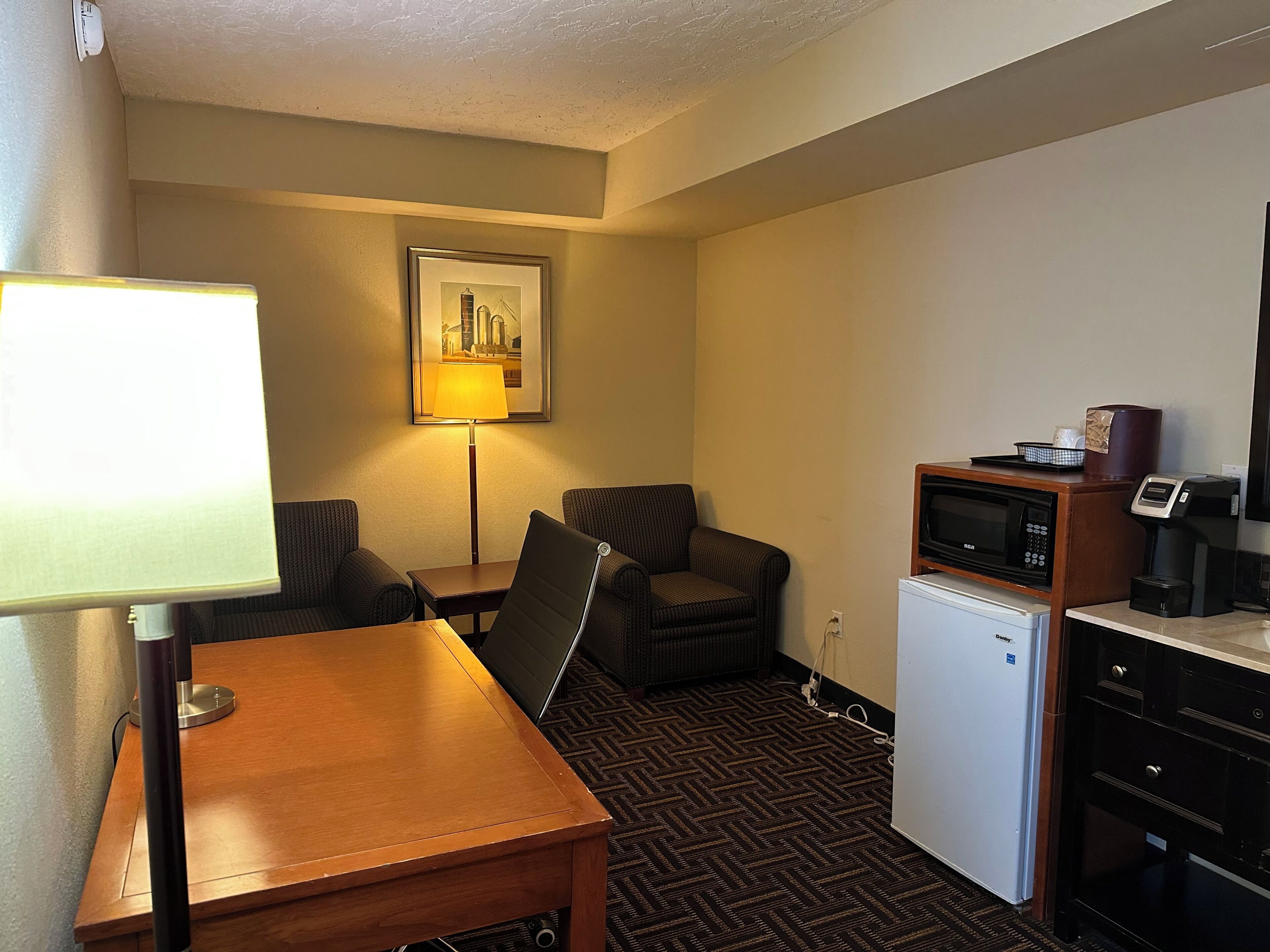 Une chambre d’hôtel compacte comprenant un bureau en bois, une chaise, un petit réfrigérateur, un four à micro-ondes, une machine à café et un tableau encadré au mur.