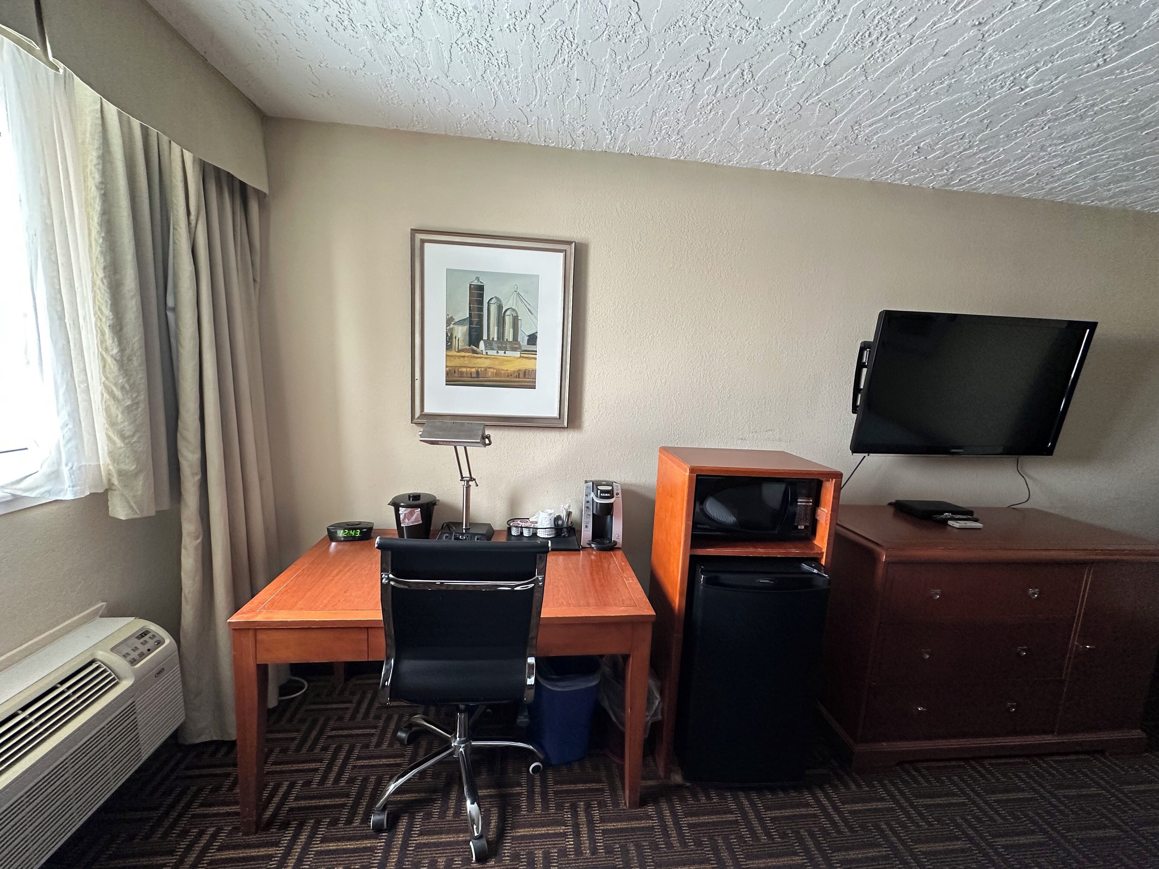 Una habitación de hotel con un escritorio de madera, una silla de oficina negra, un televisor sobre un mueble de madera, un cuadro enmarcado en la pared y una ventana con cortinas.