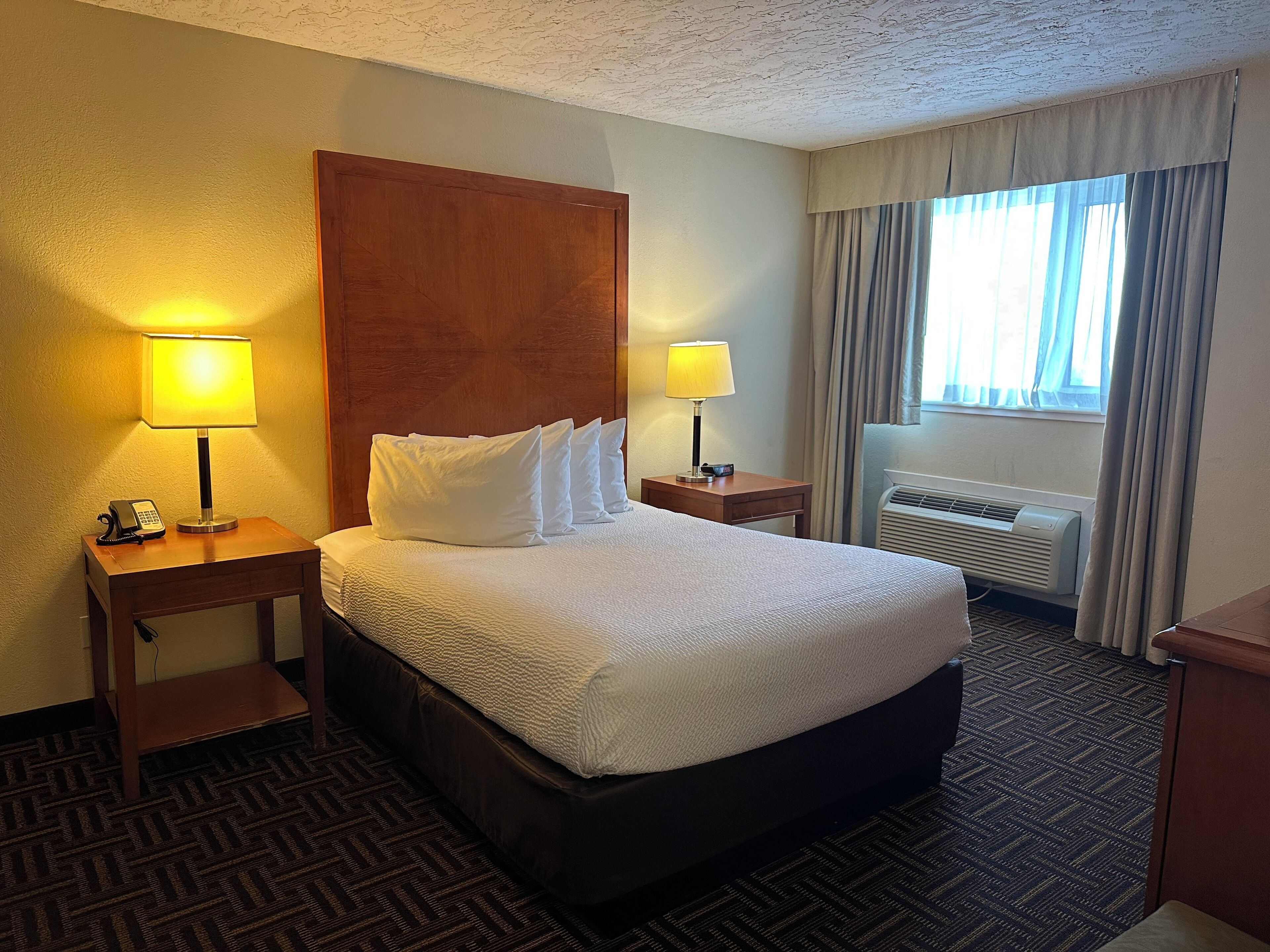 A hotel room with a large bed, two bedside tables with lamps, a telephone, and an air conditioning unit.
