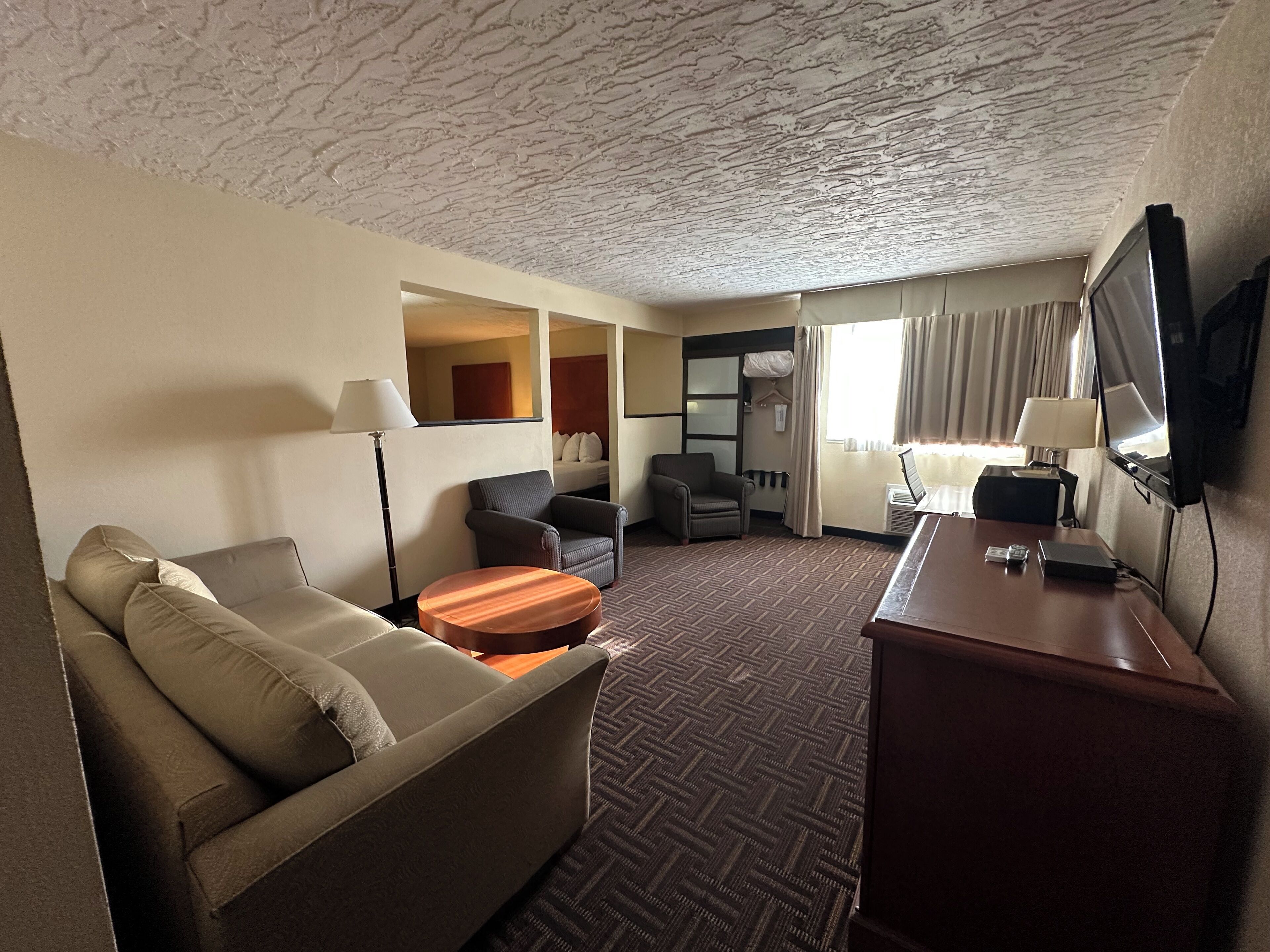 A hotel room with a sofa, armchair, small round table, desk with a TV, and a bed in the background.