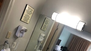Un baño de hotel con lavamanos, espejo y barra para toallas. Hay un cartel en la pared y un secador de pelo en la encimera.