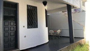 Ein überdachter Außenbereich mit einer schwarzen Tür, einem Fenster mit Metallgitter und zwei weißen Stühlen.