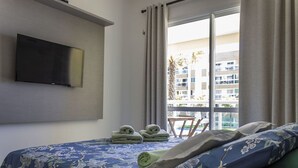 Habitación de hotel con cama, televisor de pantalla plana montado en la pared, ventana con cortinas y vista a un edificio.