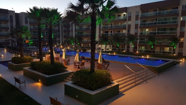 Zona de piscina hotelera con múltiples sillas de descanso y sombrillas, rodeada de palmeras y un edificio de varios pisos con balcones.
