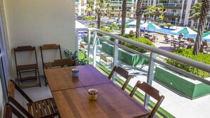 Una mesa de madera con sillas en un balcón con vistas a una zona de piscina con sombrillas y palmeras.