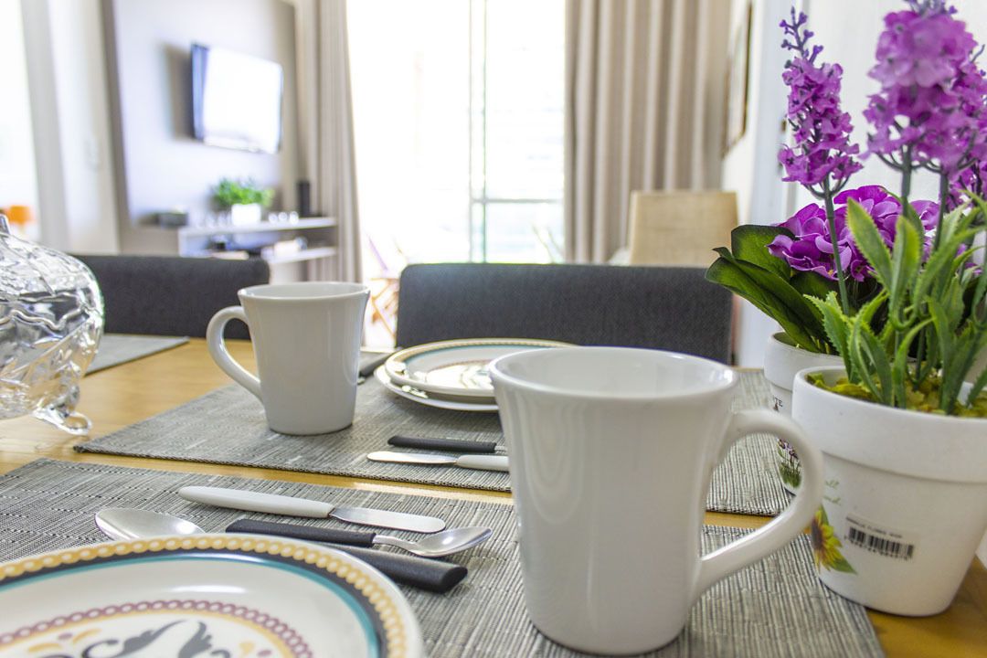 Una mesa de comedor dispuesta con tazas blancas, platos y cubiertos, una planta en maceta con flores moradas y un florero de vidrio transparente.