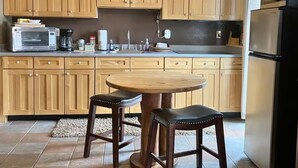 Een keuken met houten kastjes, een magnetron, een koelkast en een ronde houten tafel met twee krukjes.