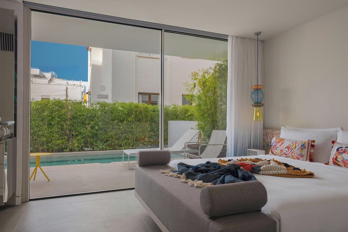 Habitación de hotel moderna con un amplio ventanal corredero que da a una zona de piscina, una cama con una manta colorida y una mesita de noche.