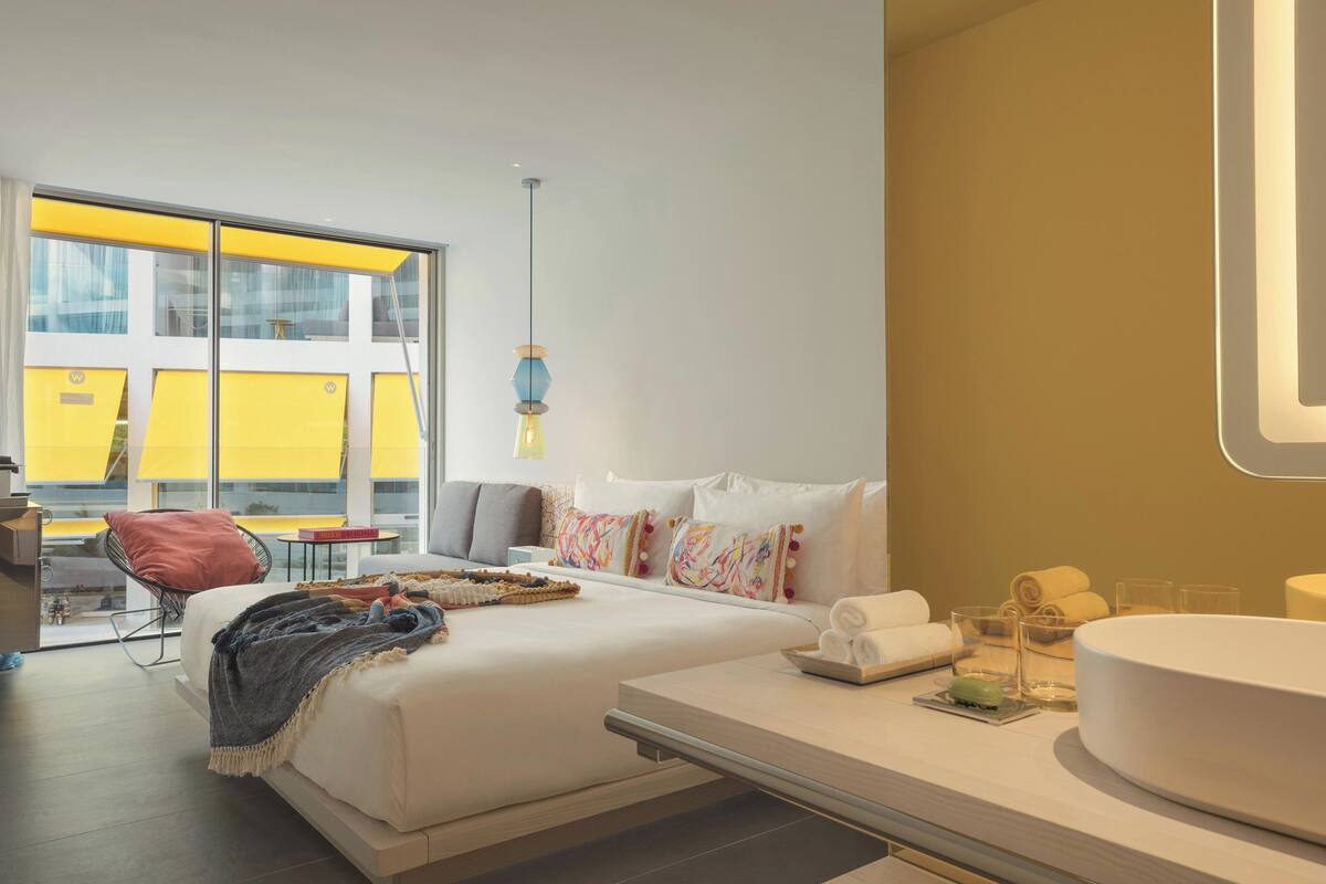 Habitación de hotel moderna con una cama grande, una mesita de noche con artículos de aseo, vistas de la ciudad a través de amplios ventanales y una pared de color amarillo brillante.