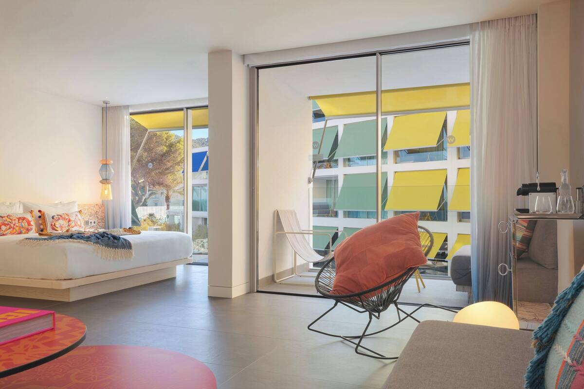 Una habitación de hotel moderna con una cama grande, un área de descanso con una silla, una mesita y vistas a un edificio con toldos coloridos.