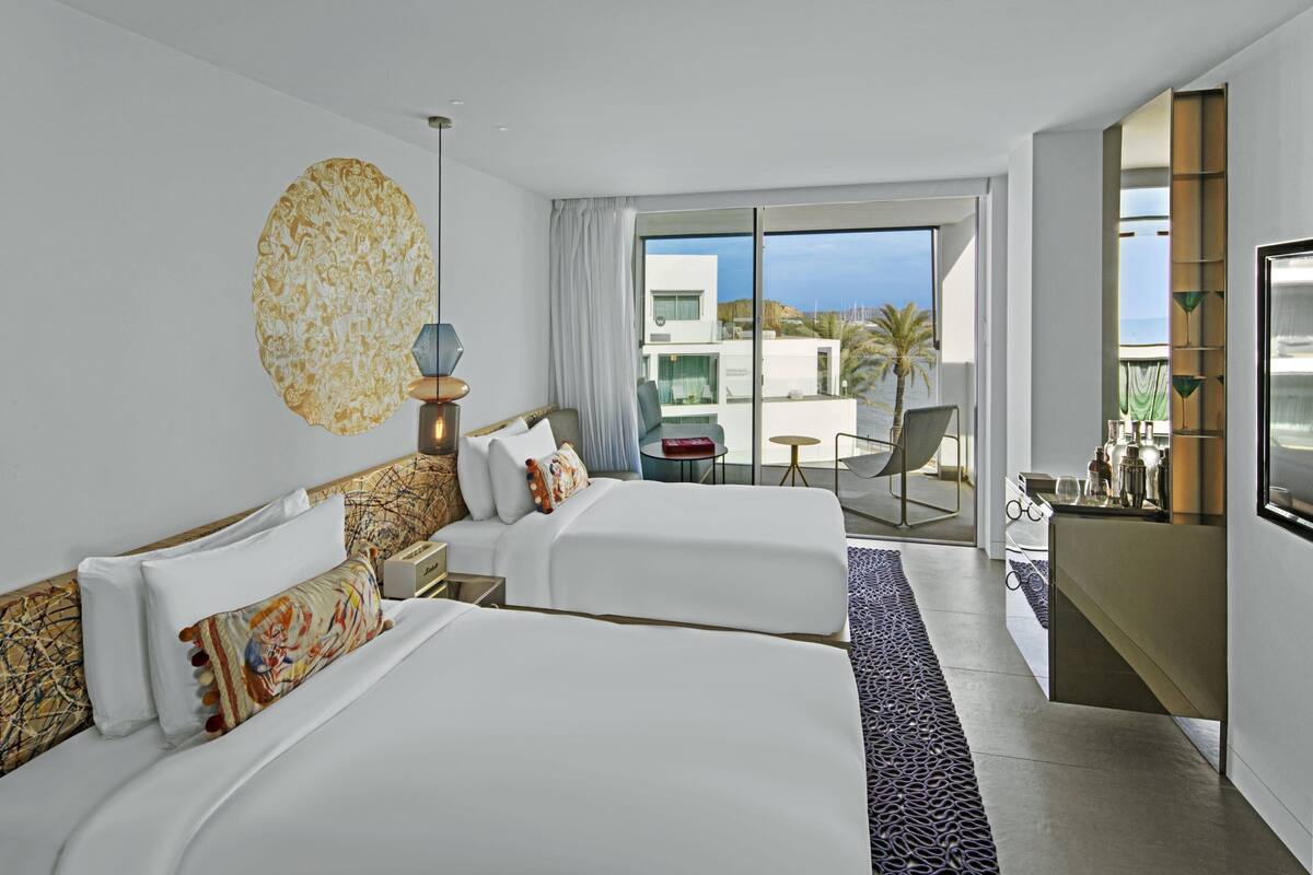 Habitación de hotel con dos camas, un balcón con vistas, un escritorio con silla y un televisor.