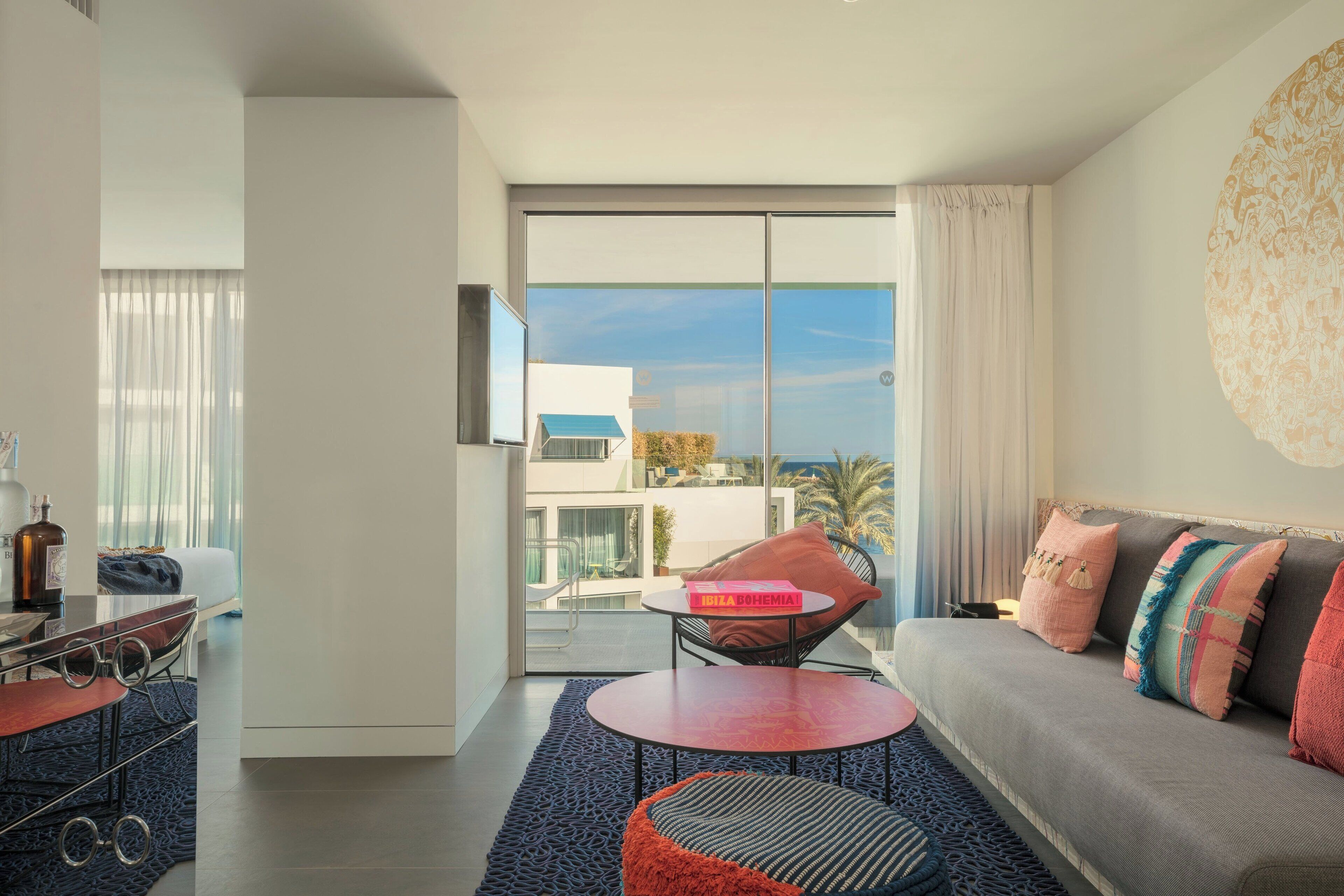 Una sala de estar moderna con un sofá, una mesa de centro y una silla. Hay un ventanal con vistas a edificios y palmeras.