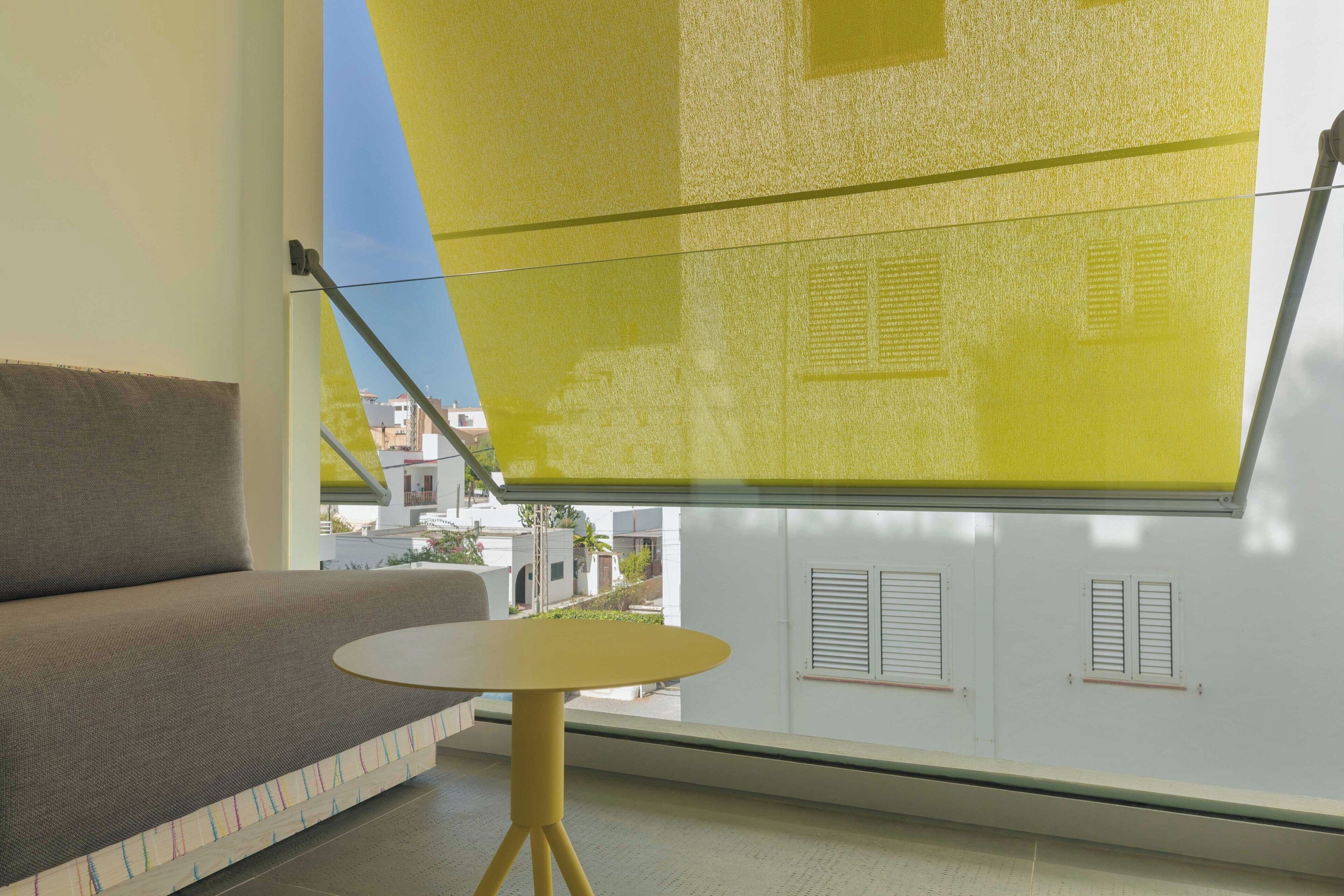 Quarto com sofá, mesa redonda pequena, persiana amarela e vista para edifícios.