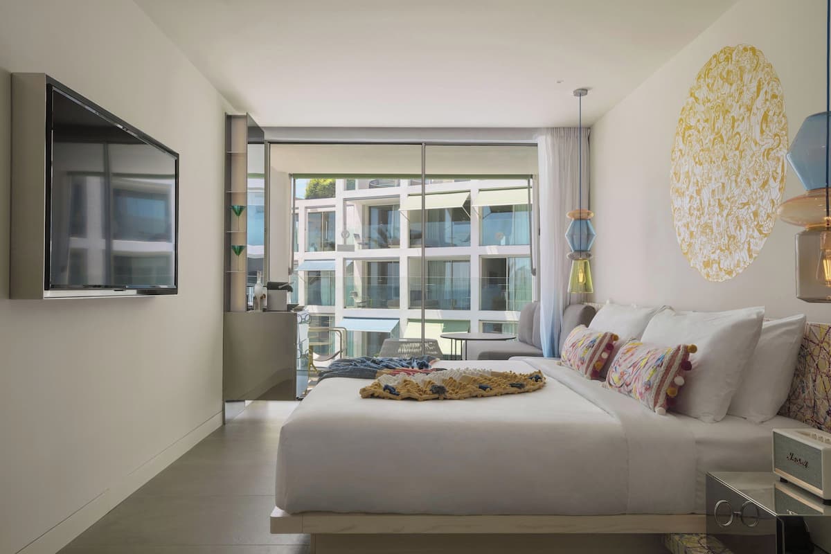 Habitación de hotel moderna con una cama grande, un televisor de pantalla plana en la pared, un ventanal con vistas al edificio y una pieza decorativa en la pared.