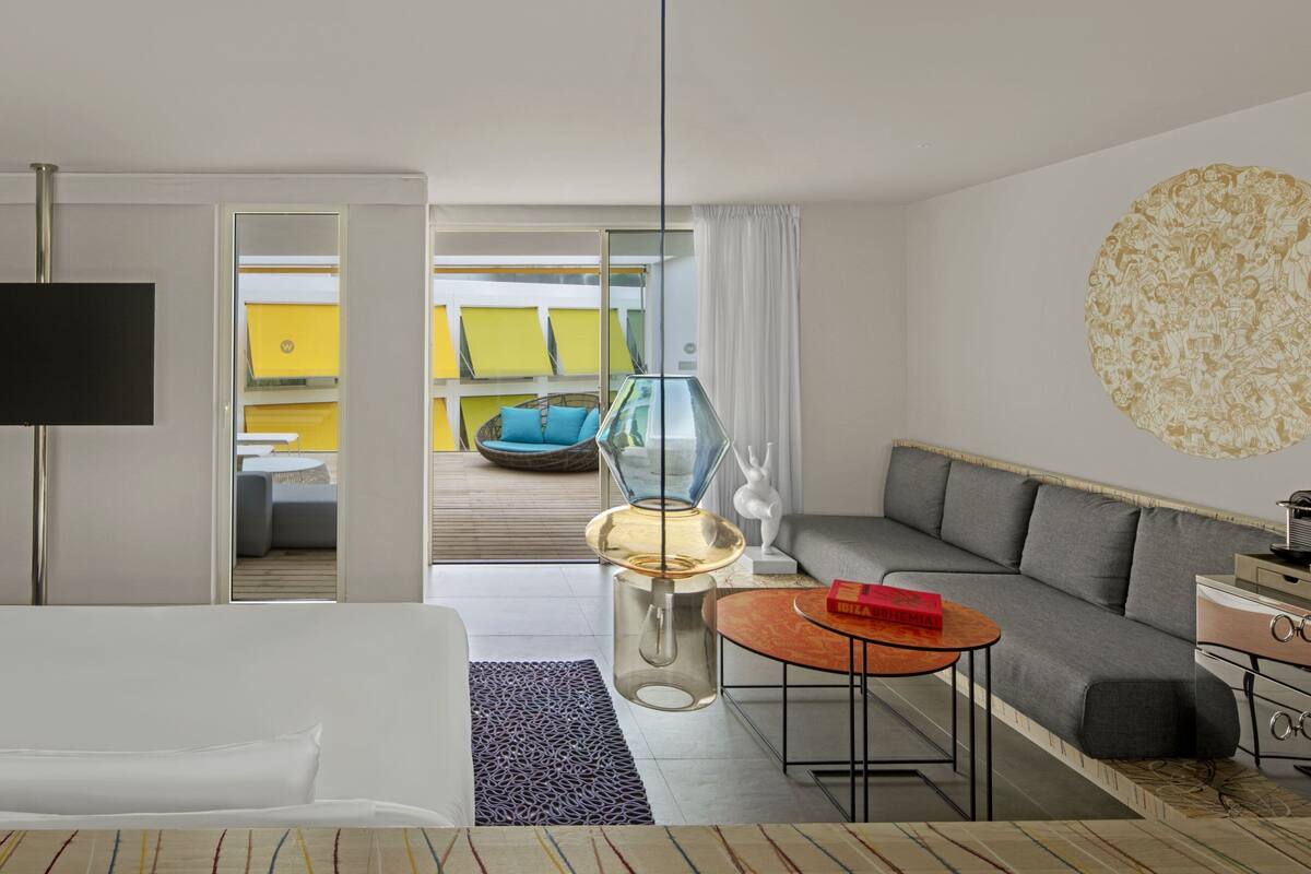 Una habitación de hotel moderna con un sofá, una mesita redonda pequeña, un televisor y un separador de vidrio que da a un balcón con una silla azul.