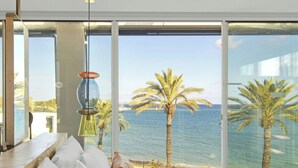 Quarto com uma grande janela com vista para uma praia e o mar. Tem uma cama com roupa de cama branca e um travesseiro colorido.