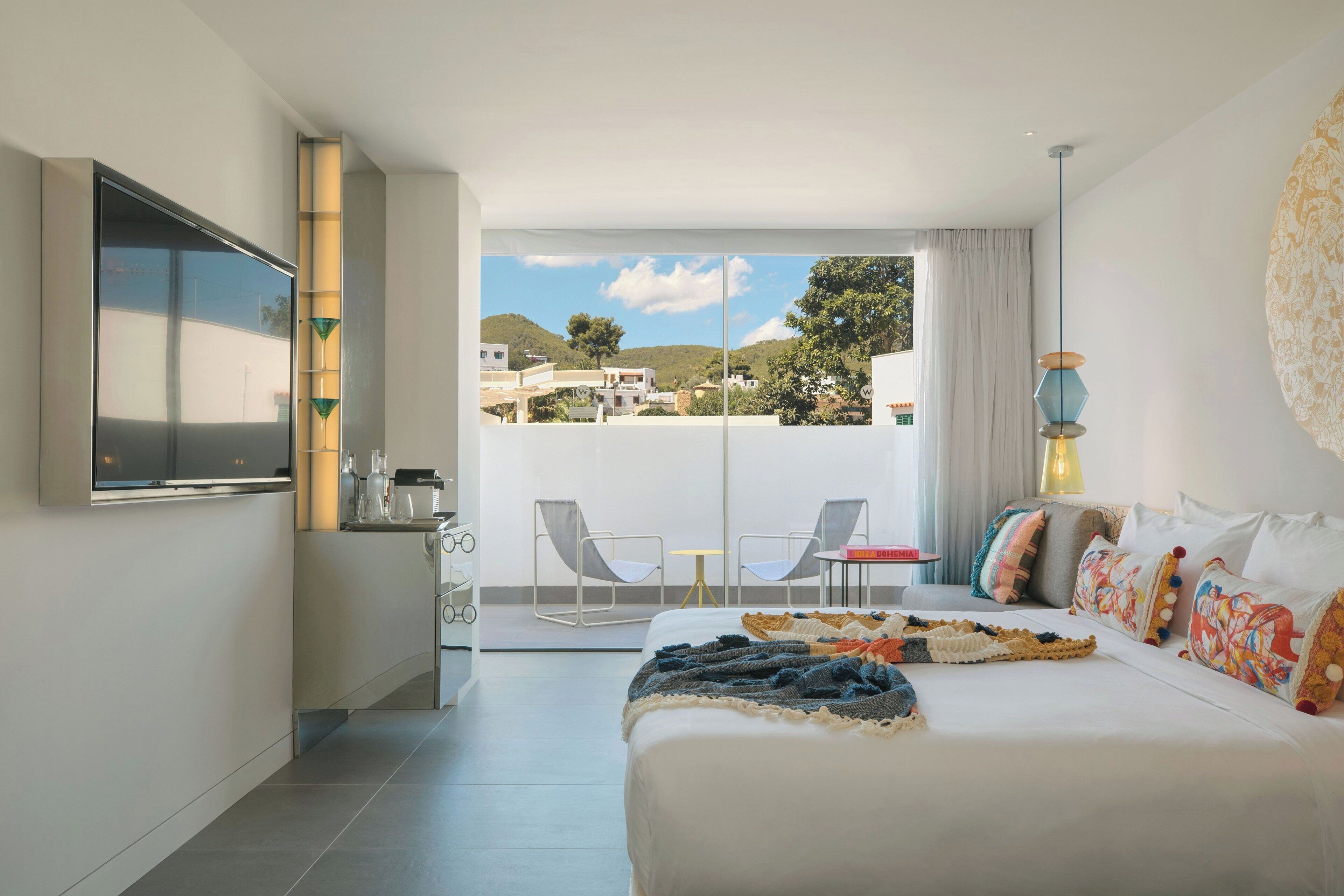 Una habitación de hotel moderna con una cama grande, un televisor de pantalla plana, un balcón con sillas y vistas del entorno.