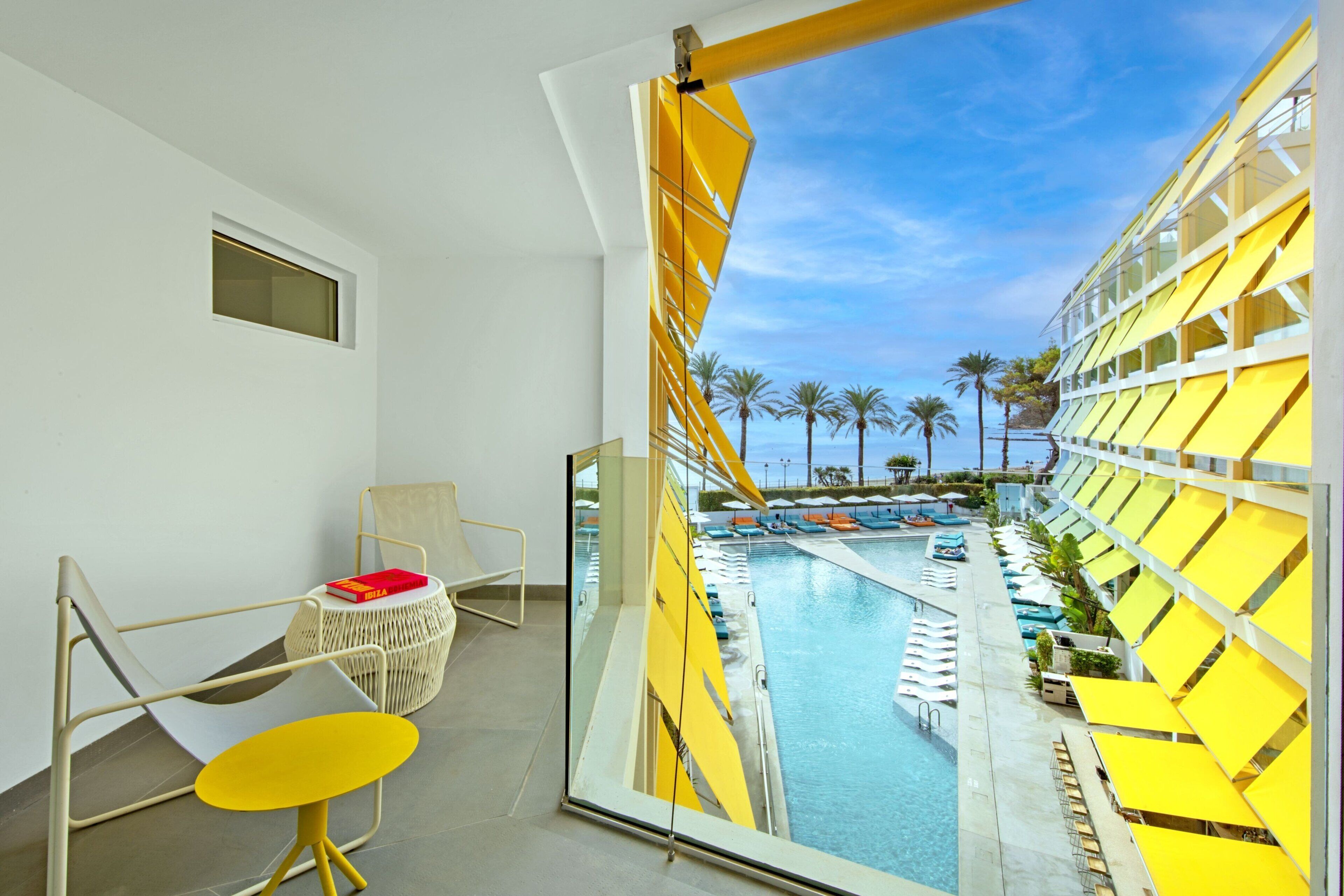 A hotel room with a balcony overlooking a pool area, featuring yellow sun loungers and palm trees.