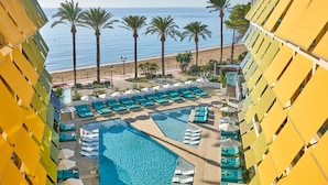Uma área de piscina de hotel com vários espreguiçadeiras e guarda-sóis, uma grande piscina e vista para a praia com palmeiras.