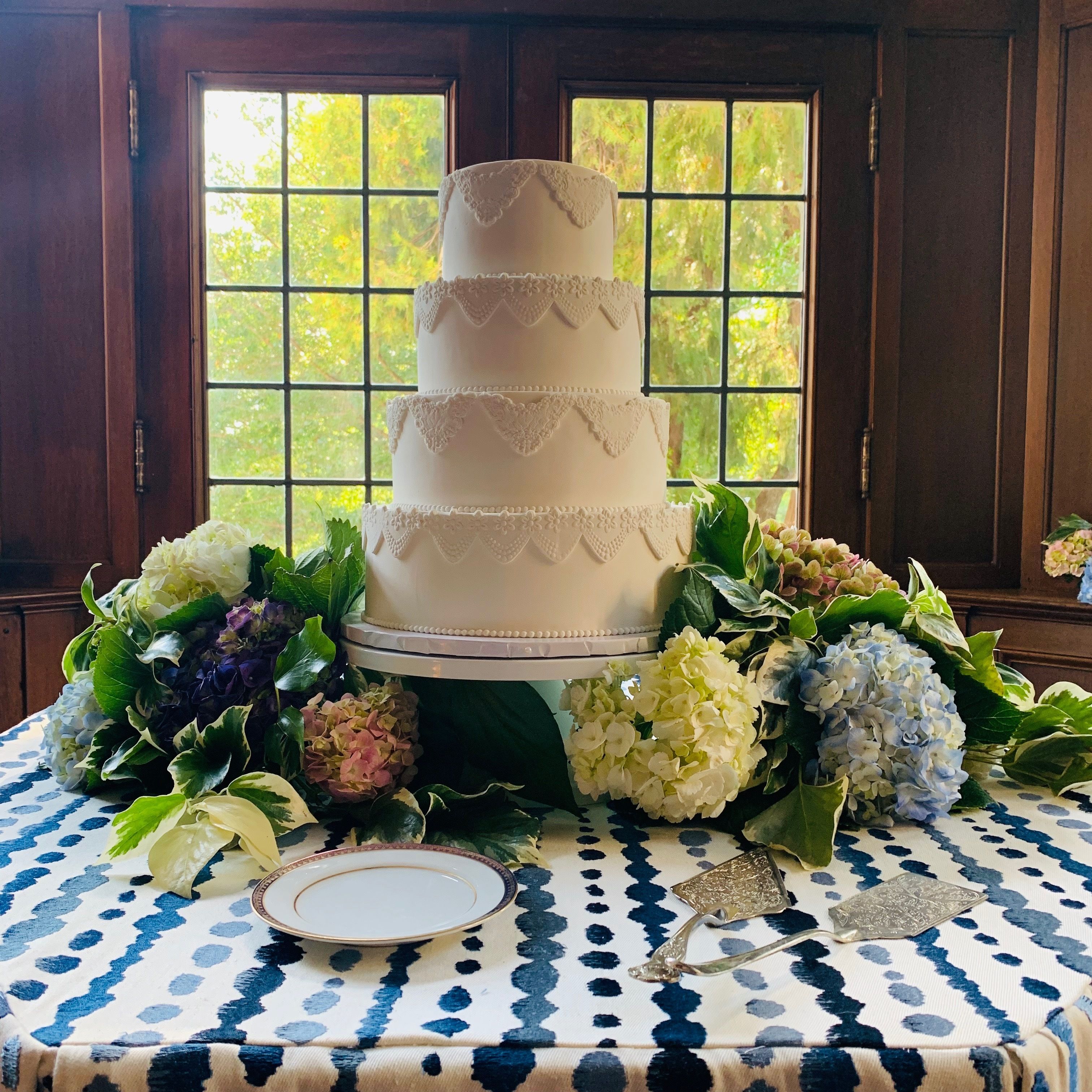 A multi-tiered wedding cake with lace details, surrounded by floral arrangements on a patterned tablecloth.