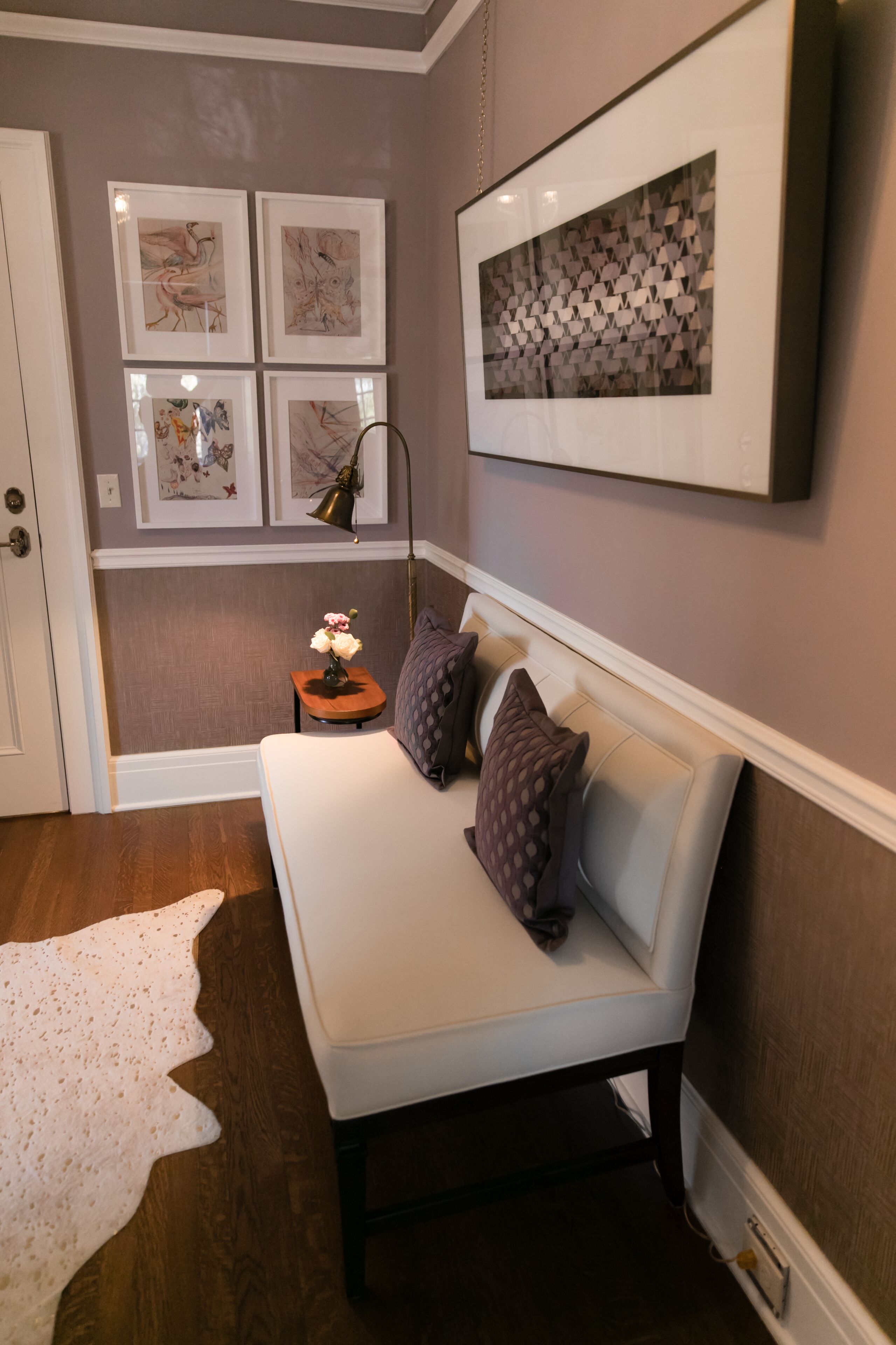 A room with a white sofa, a small side table with a lamp, and a white rug. There are four framed artworks on the wall.
