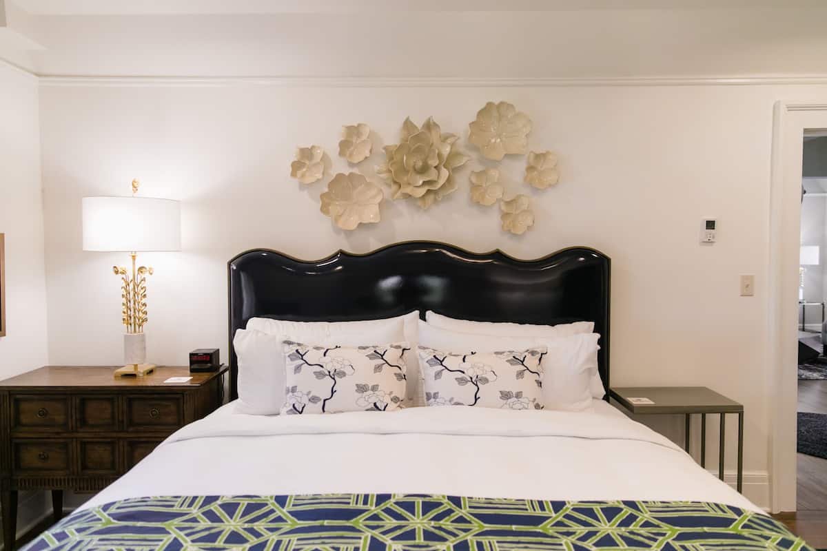 A neatly made bed with a patterned pillow, a wall-mounted decorative piece, a bedside table with a lamp, and a small nightstand.