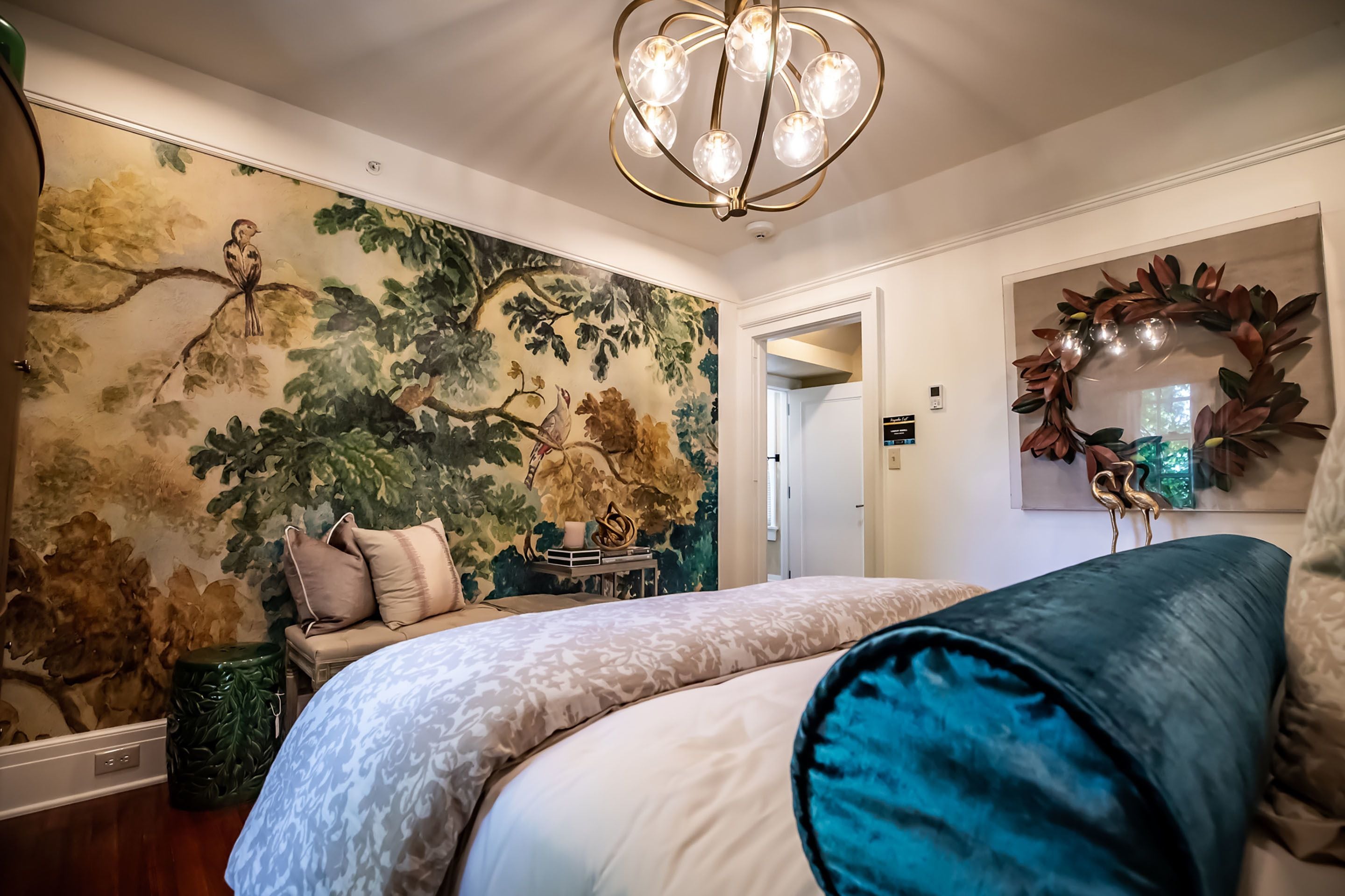Een slaapkamer met een groot bed, een muurschildering van vogels, een kroonluchter en een spiegel met een decoratieve krans.