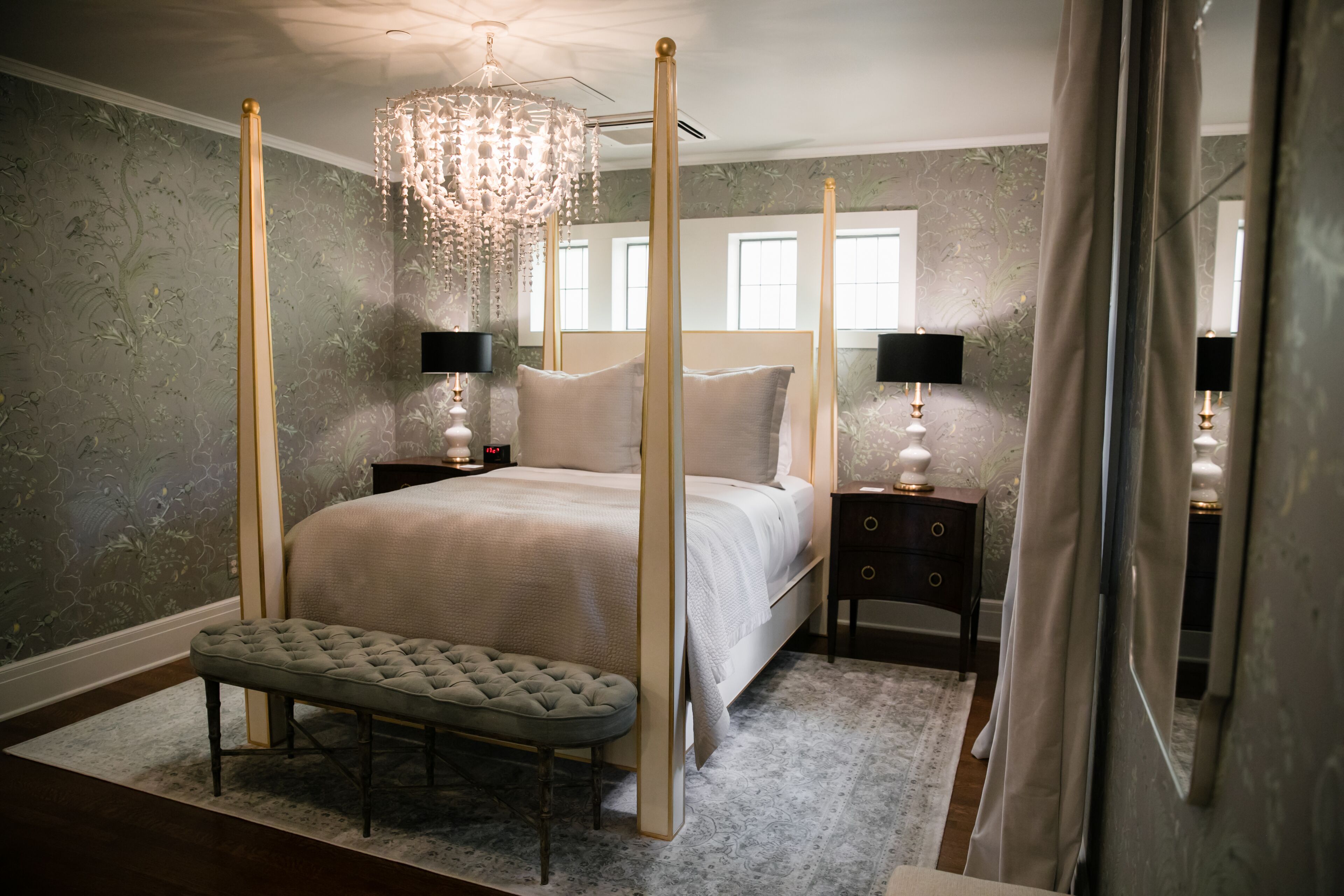 A four-poster bed with a tufted bench, bedside tables with lamps, and a chandelier.