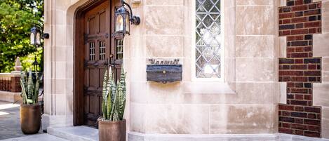 A building entrance with a wooden door, a window with a diamond pattern, and two potted plants.