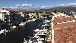 Eine Marina mit vertäuten Booten entlang eines schmalen Wasserwegs, umgeben von Gebäuden mit roten Ziegeldächern und einer Bergkulisse.