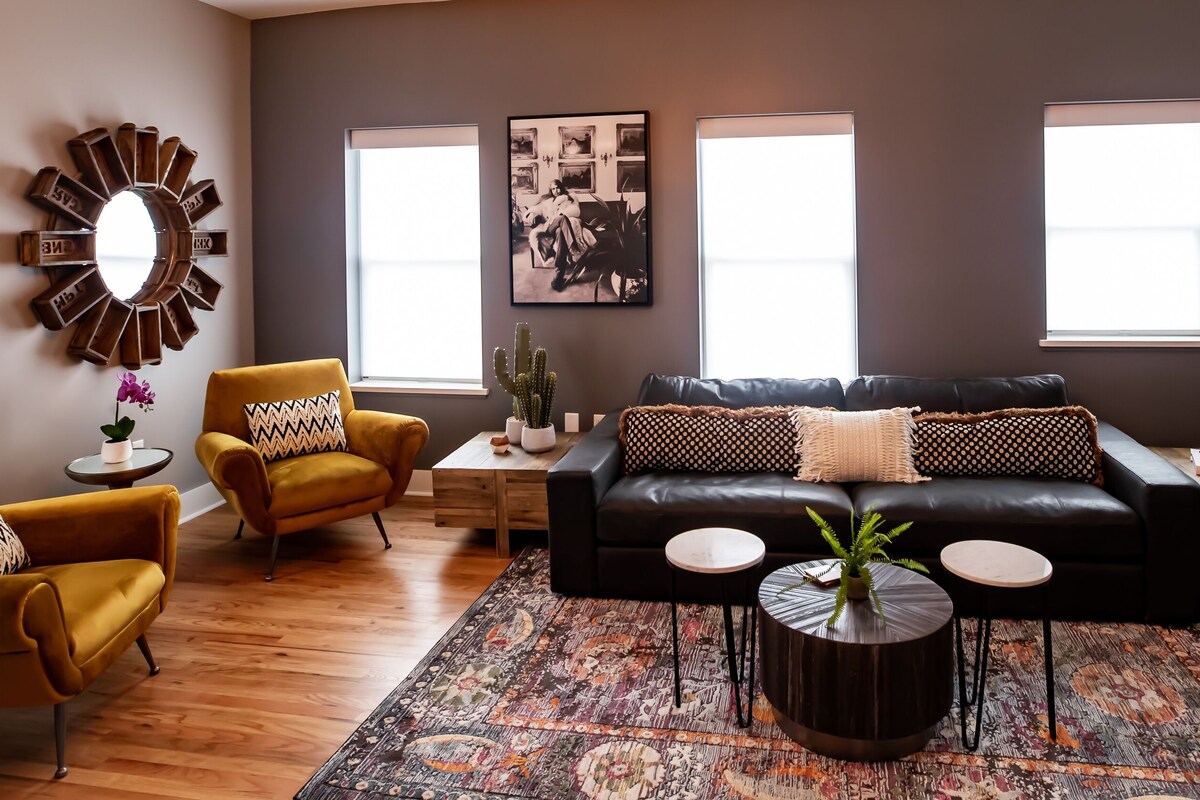 A modern living room with a black leather sofa, mustard armchairs, a patterned rug, and a large mirror on the wall.